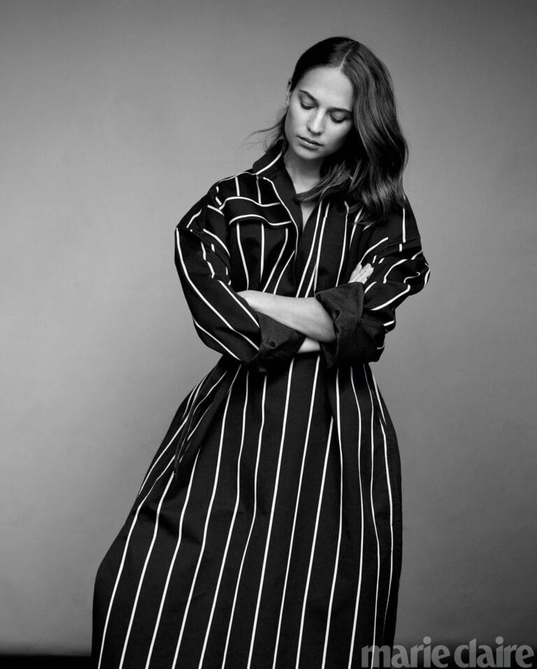 Alicia Vikander: Wears a long white-striped shirt over a black background, with a wide cut…