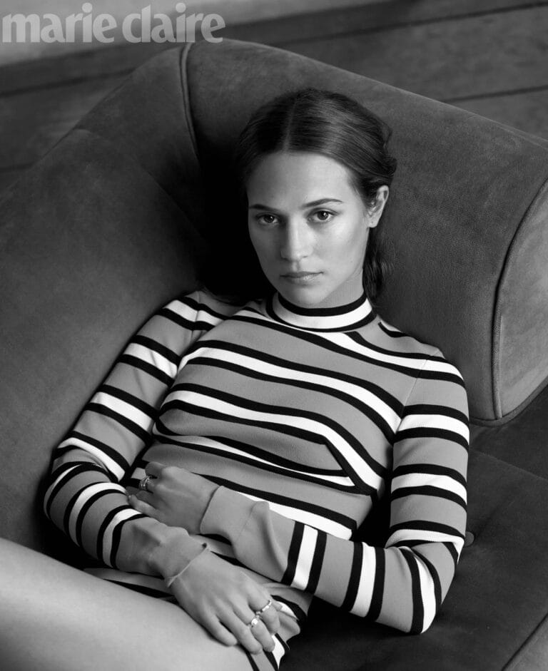 Alicia Vikander: Poses reclined on a velvet sofa, wearing a striped sweater in shades of…