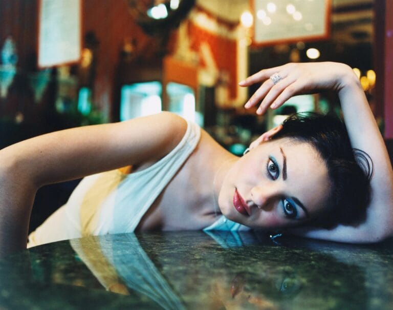 Mila Kunis: Wears a white top with thin straps, leaning on a marble table with…