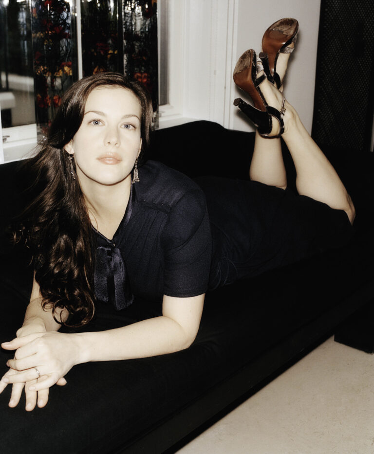 Liv Tyler: Wearing a black dress with an elegant bow, reclining on a dark sofa while…