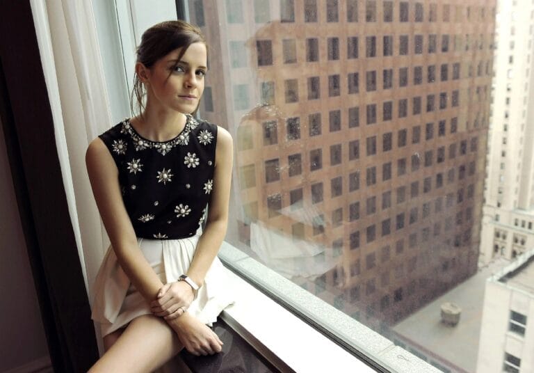 Emma Watson: Wears a black blouse with shiny details and a white pleated skirt, sitting next…