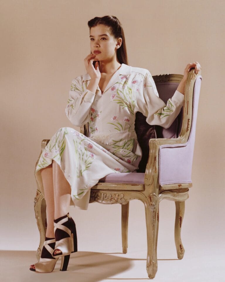 Hailee Steinfeld: Wears a long white silk dress with delicate floral prints, while sitting…