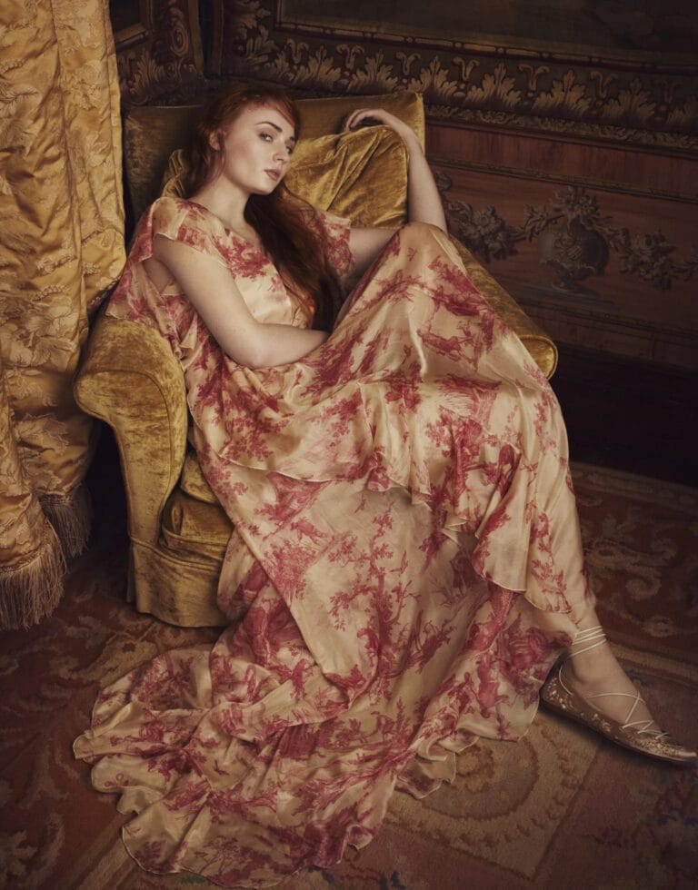 Sophie Turner: Wears a flowy cream dress with red floral prints, sitting…