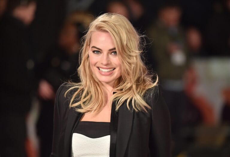 Margot Robbie: Wears a black jacket and set with a textured white top, smiling confidently…