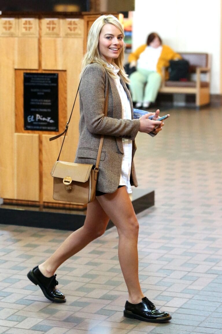 Margot Robbie: Camina con confianza en un entorno urbano, luciendo un blazer marrón sobre una blusa…