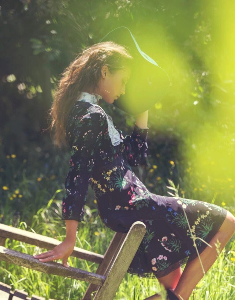 Alicia Vikander: Wears a black dress with a floral print, seated on a wooden structure, with…