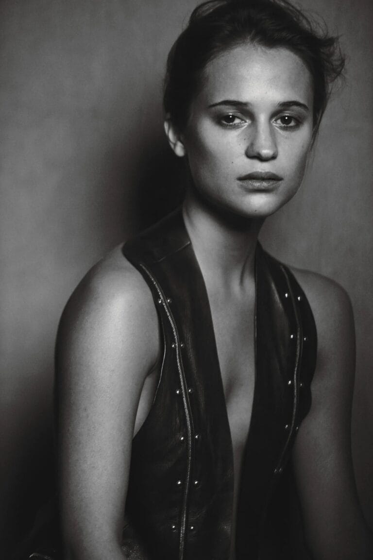 Alicia Vikander: Wears a black leather piece with a deep neckline and metallic details, seated…