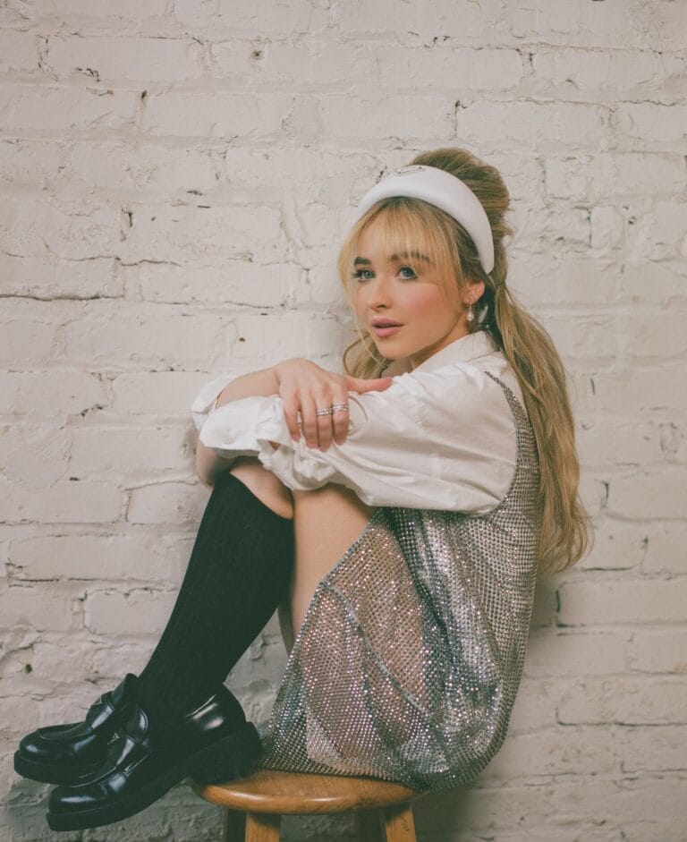 Sabrina Carpenter: Está sentada en un taburete de madera, abrazando sus piernas, con una blusa blanca…