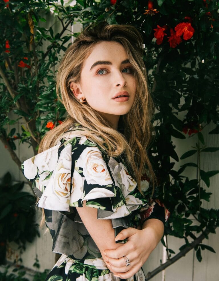 Sabrina Carpenter: Posa entre un fondo de plantas verdes y flores rojas, luciendo un vestido de…