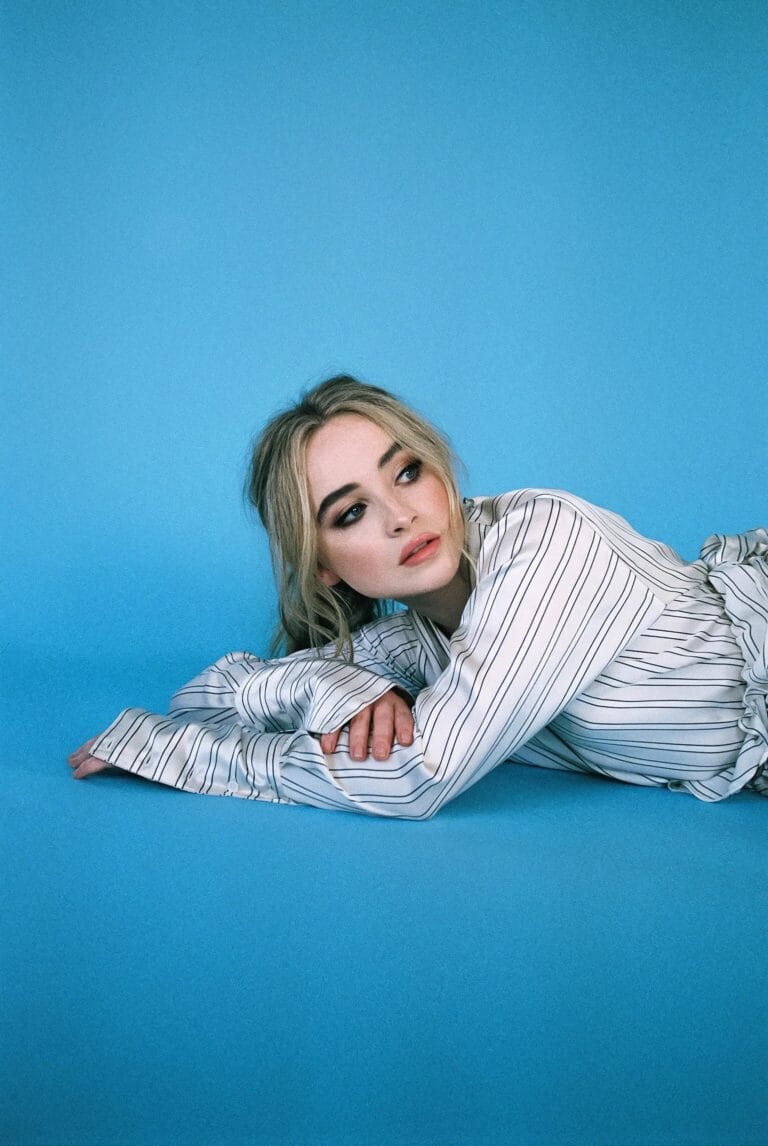 Sabrina Carpenter: Aparece recostada sobre un fondo azul, vistiendo una blusa de seda blanca con rayas…