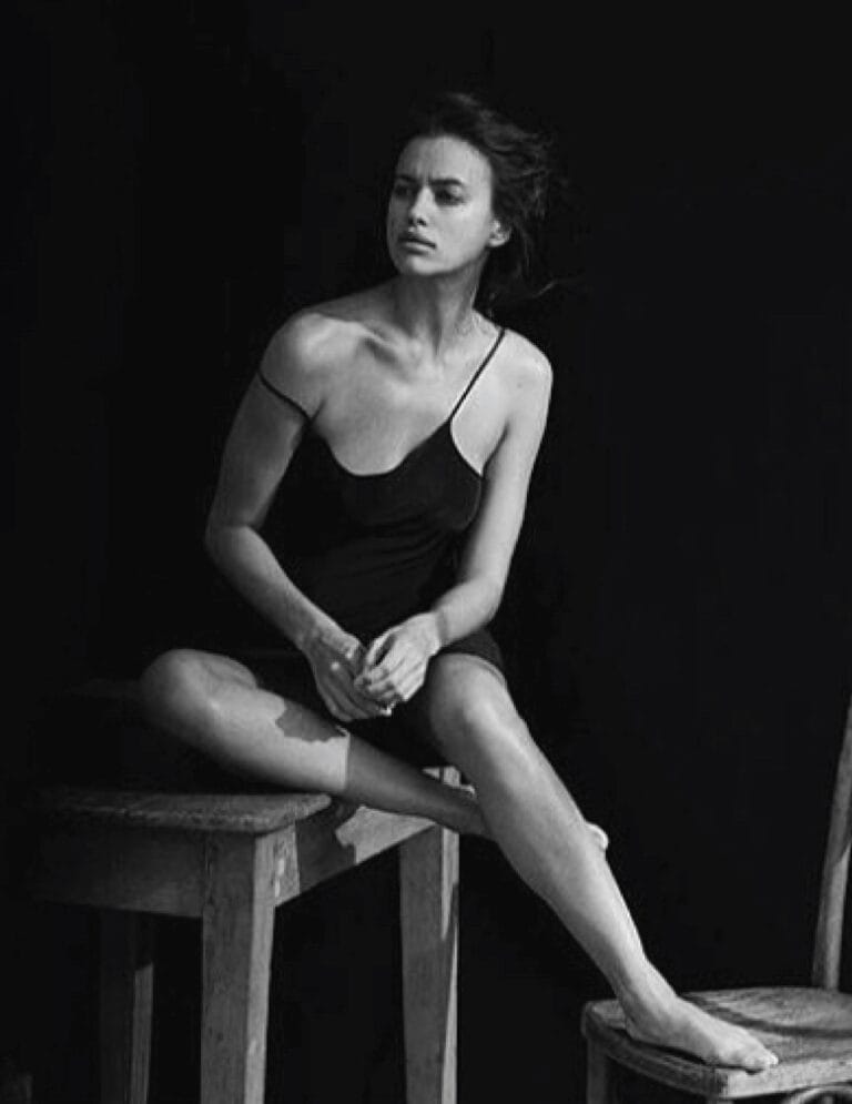 Irina Shayk: Wears a black slip dress with thin straps, sitting on a stool with one leg…