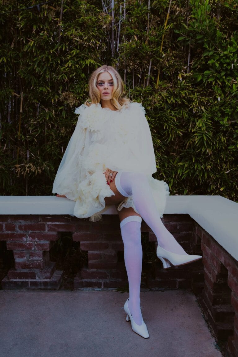 Samara Weaving: Sits on a bench in a white dress with ruffled details that…