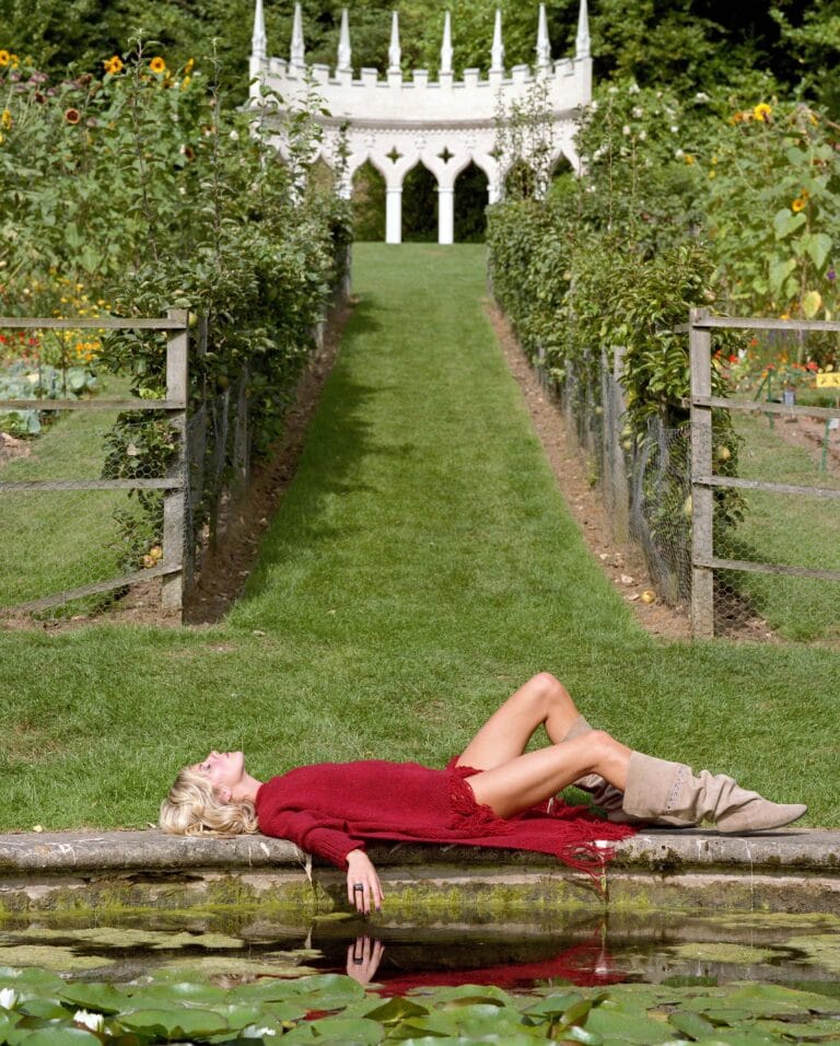 Sienna Miller: Lies on a stone by a pond, wearing a red sweater with…