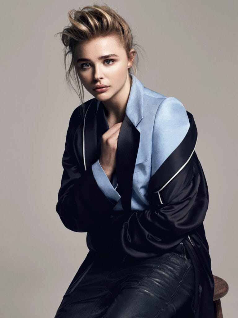 Chloë Grace Moretz: Wears a light blue satin blouse under a black coat with white details,…