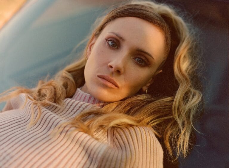 Juno Temple: Wears a striped sweater in soft tones, lying on a surface with a…