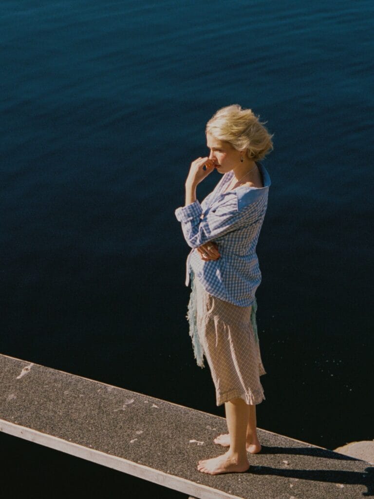 Thea Sofie Loch Næss: Poses on a pier, barefoot, in a blue and white plaid shirt that…