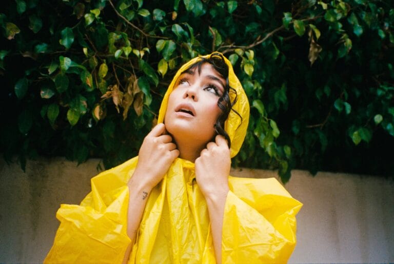 Courtney Eaton: Luce un impermeable amarillo brillante, con la capucha puesta, mientras sostiene el tejido cerca…