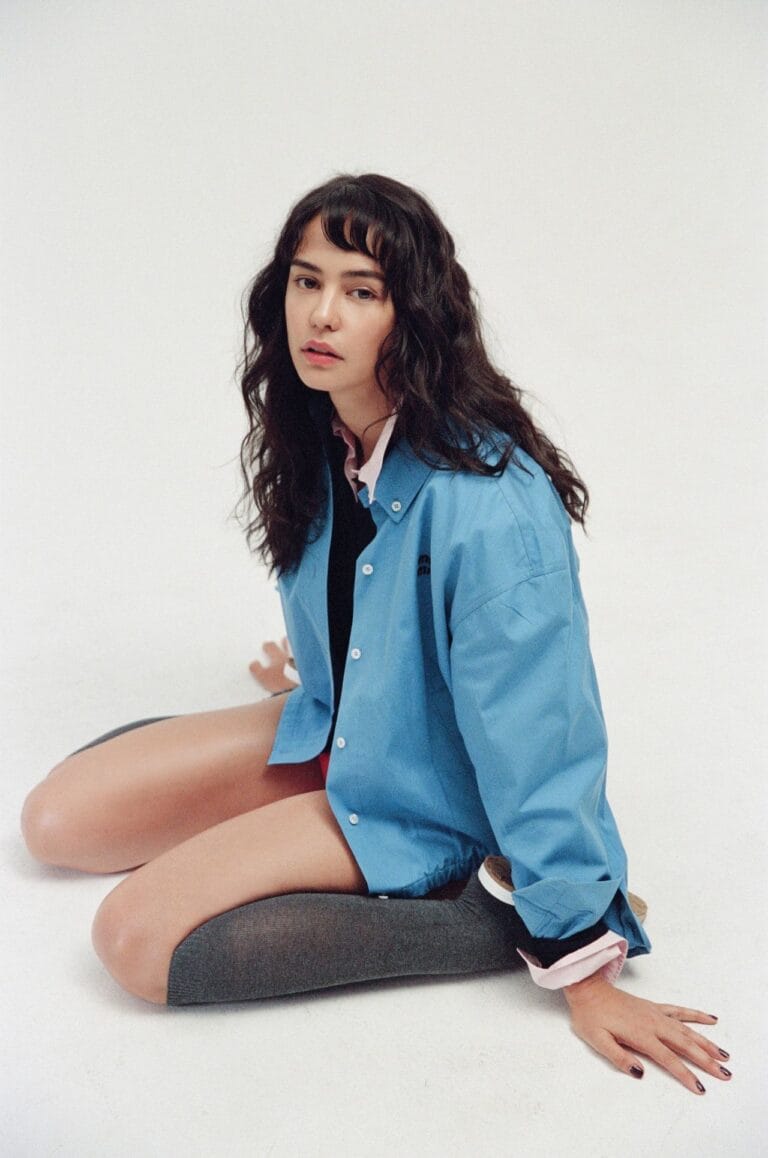 Courtney Eaton: Luce una camisa azul oversized sobre una blusa rosa, sentada en el suelo con…