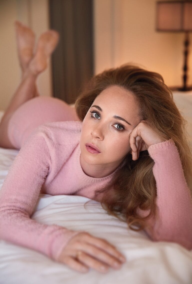 Juno Temple: Está tumbada sobre una cama, luciendo un suéter rosa de manga larga que abraza…