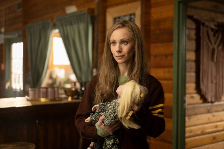 Juno Temple: Luce un suéter marrón con rayas amarillas, sosteniendo una muñeca con un vestido estampado…