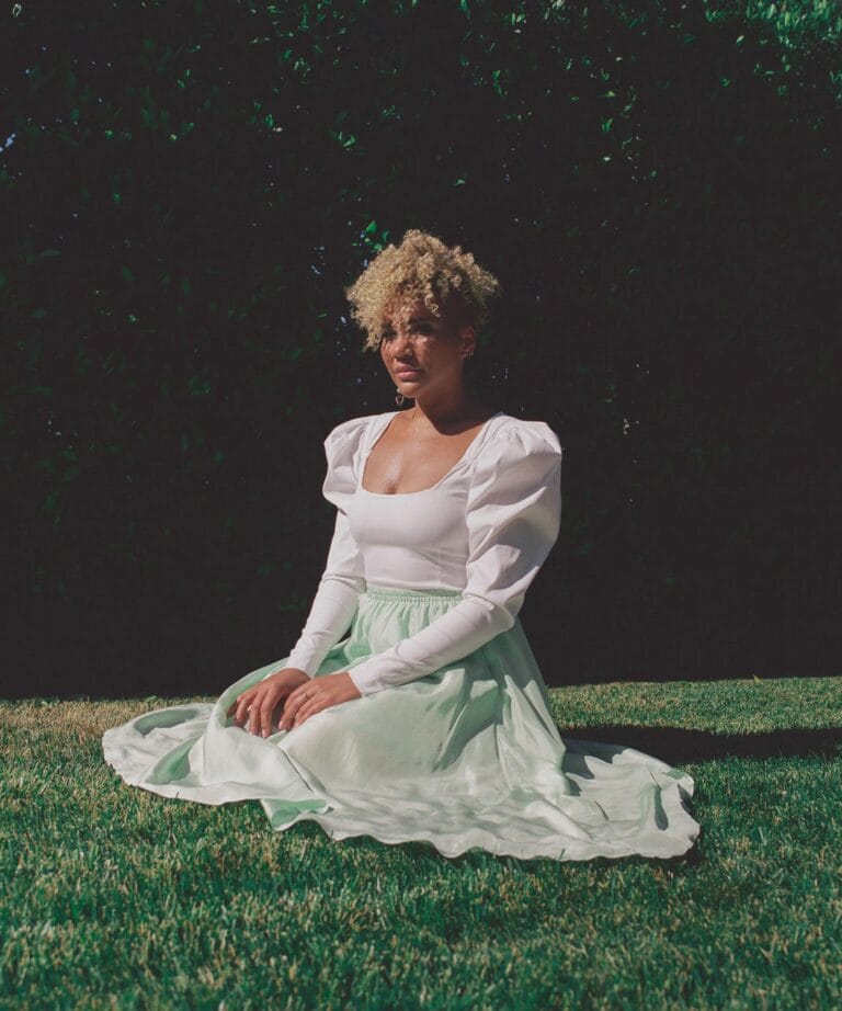 Emmy Raver-Lampman: Wears a white top with puffed sleeves and a long pastel green skirt, seated…
