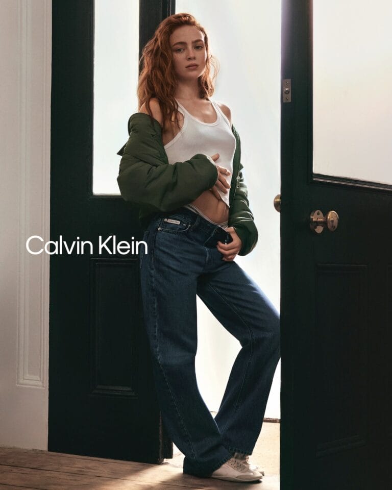 Sadie Sink: Luce una camiseta blanca ajustada bajo una chaqueta verde oliva, apoyada en el marco…