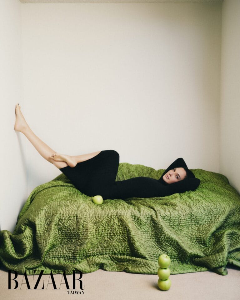 Gal Gadot: Poses against a minimalist backdrop, lying on a bed covered with a green quilt,…