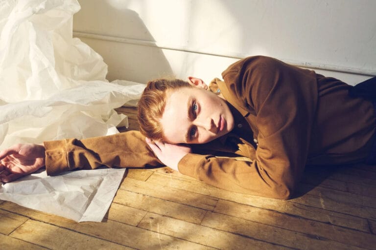 Sadie Sink: Lies stretched out on a wooden floor, in a brown outfit that stands out for…