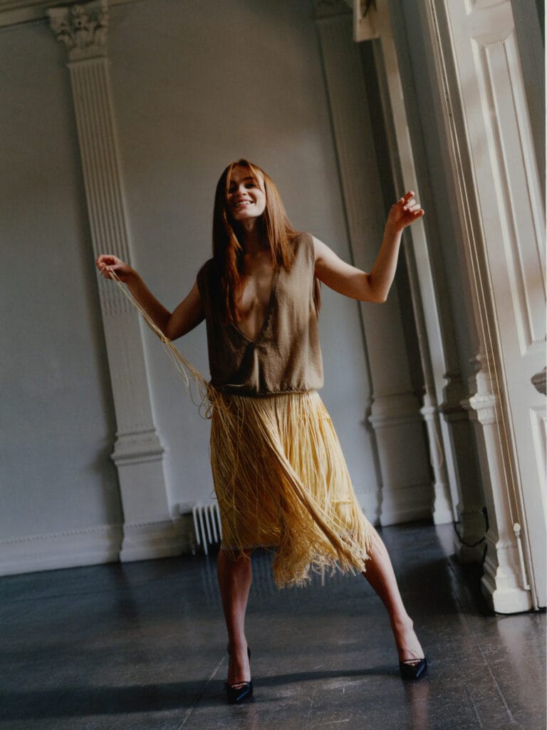 Sadie Sink: Wears a brown top with a deep neckline and a skirt with yellow fringe…