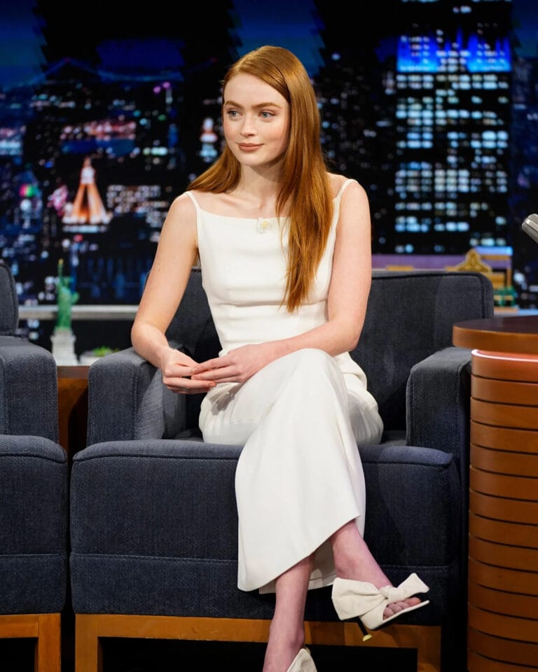 Sadie Sink: Wears an elegant white dress with thin straps, seated on a sofa with her…