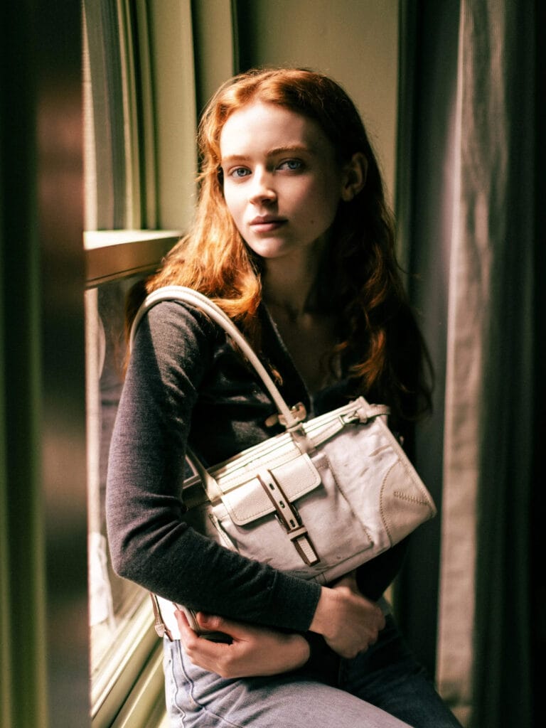 Sadie Sink: Poses by a window in a fitted gray sweater and light-wash jeans that…