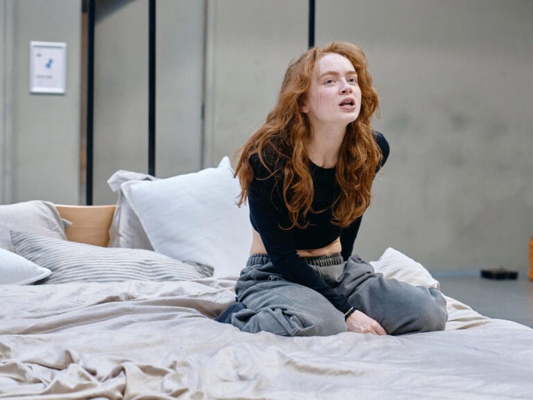 Sadie Sink: Seated on a messy bed with a introspective posture, wearing a black top…