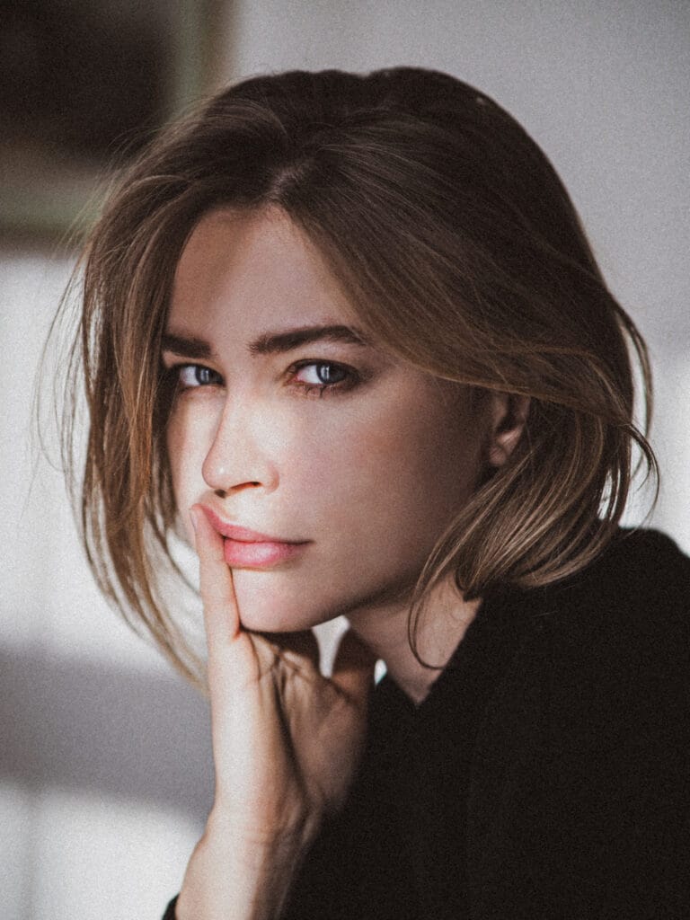 Jeanne Goursaud: Wears a black sweater, with a contemplative expression as she rests her hand on the…
