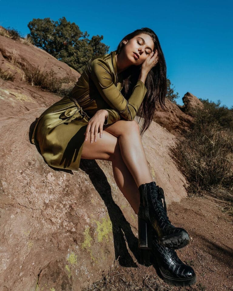 Kira Kosarin: Poses on a rock, wearing an olive-green flowy dress that highlights…