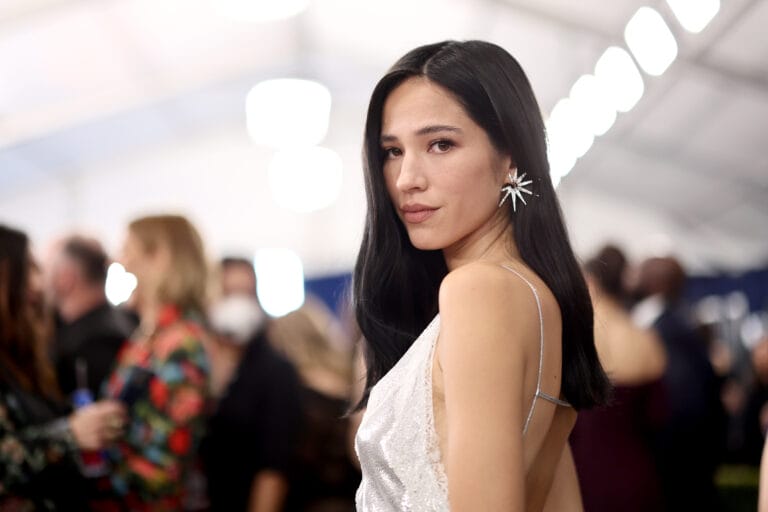 Kelsey Asbille: Wears a silver dress with thin straps, with an elegant design that highlights her…