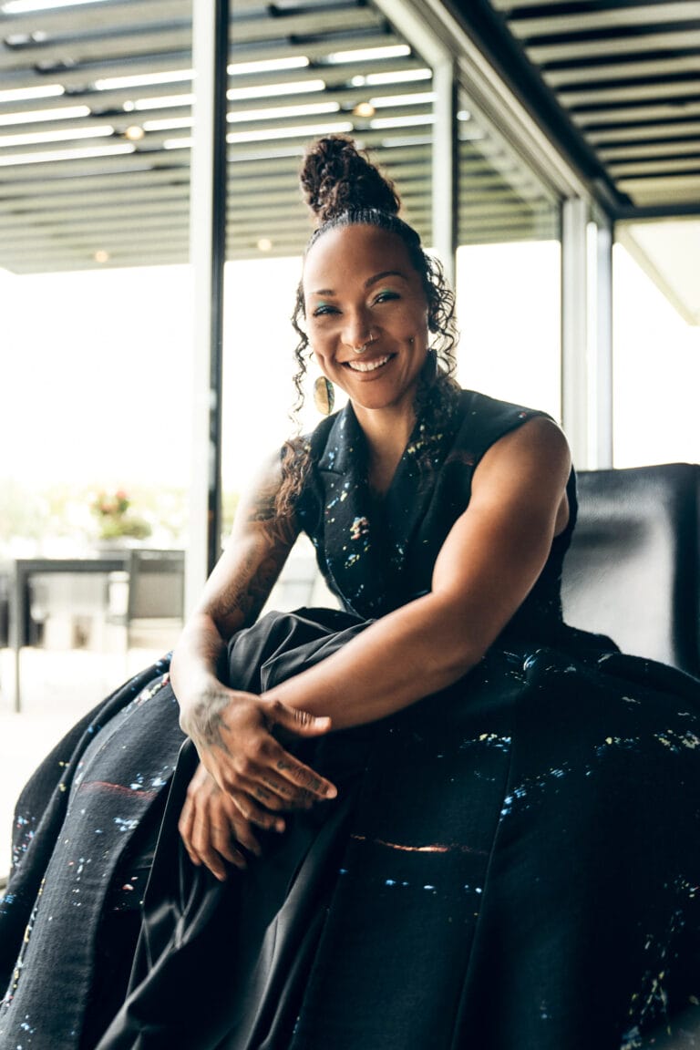 Kali Reis: Sits on a modern armchair, wearing a black dress with sparkling details that…