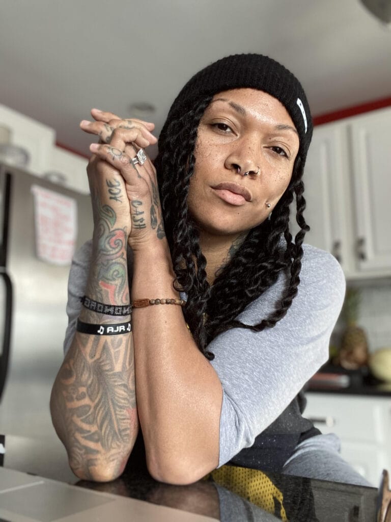 Kali Reis: Wears a gray long-sleeve T-shirt, sitting with her arms crossed over the…