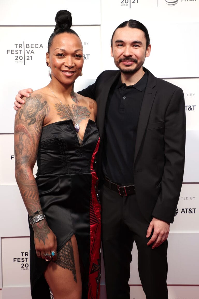 Kali Reis: Wears a black dress with an asymmetrical cut and a red detail that highlights…