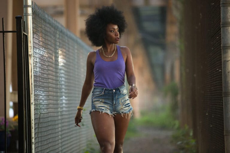 Teyonah Parris: Walks confidently down an urban hallway, wearing a purple sleeveless top and…