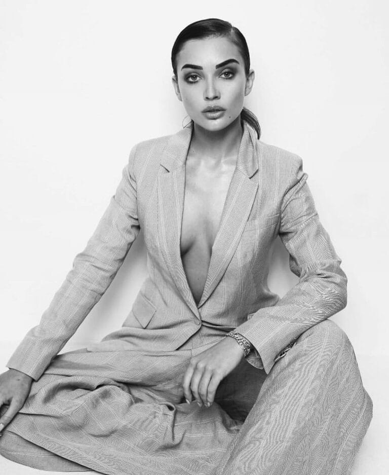 Amy Jackson: Wears a gray suit with a deep neckline, sitting on the floor…