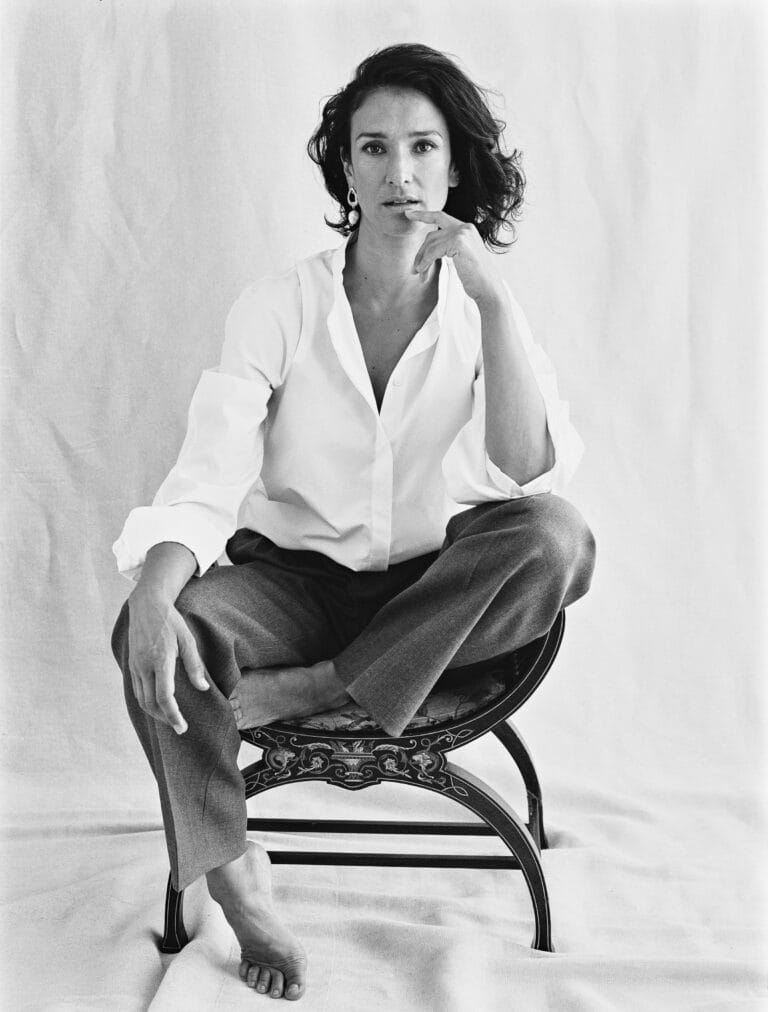 Indira Varma: Luce una camisa blanca con un escote sutil y pantalones grises, sentada en una…