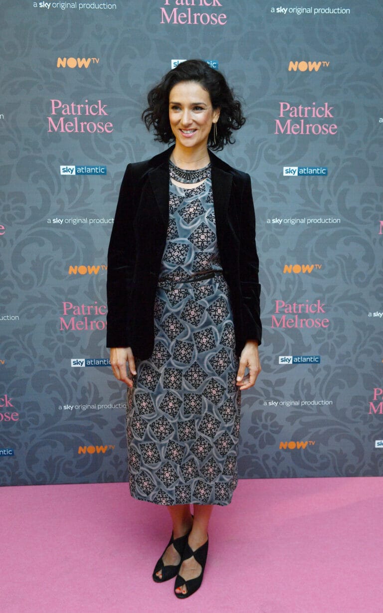 Indira Varma: Posa en un fondo decorado con motivos elegantes, vistiendo un vestido midi de estampado…