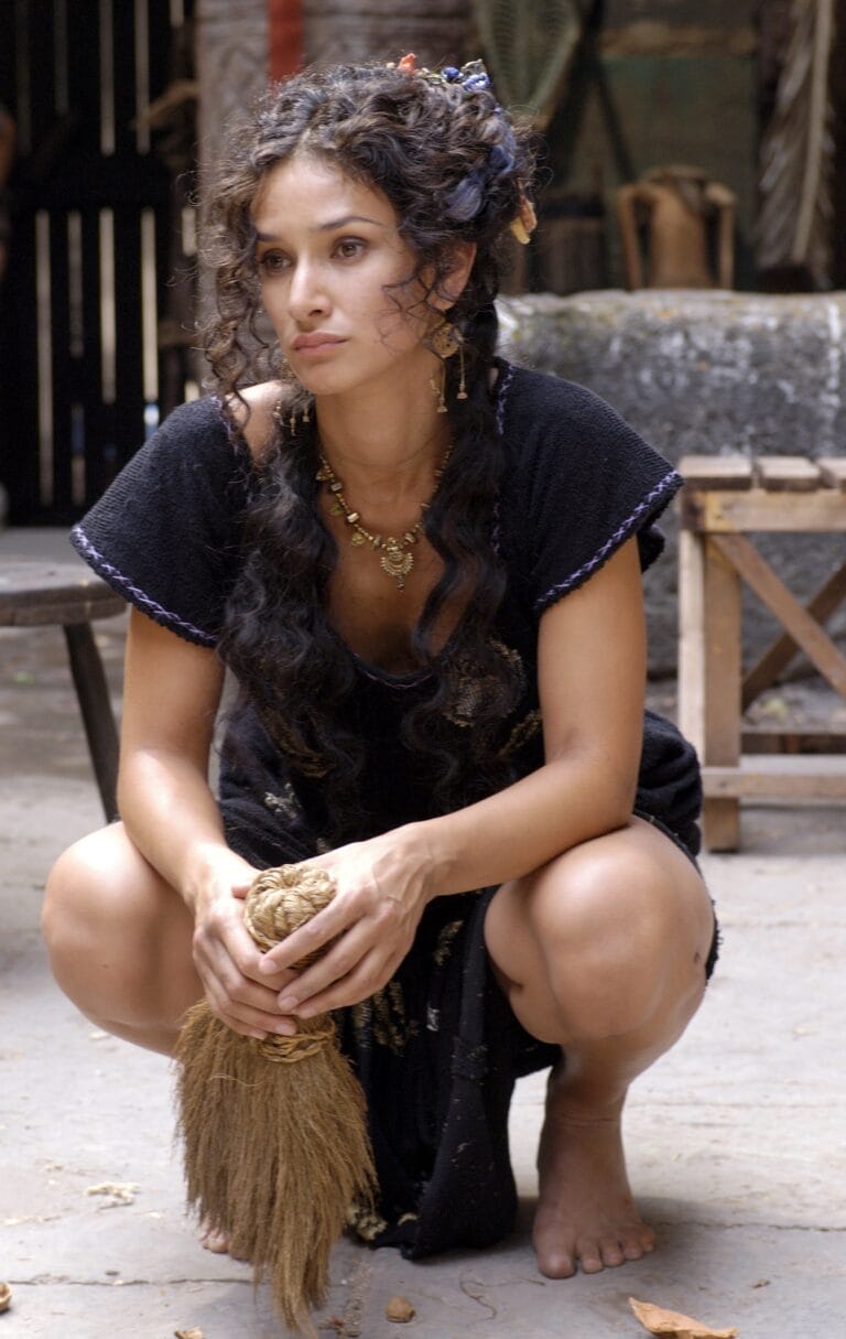 Indira Varma: Crouches barefoot on the stone floor in a black dress that…
