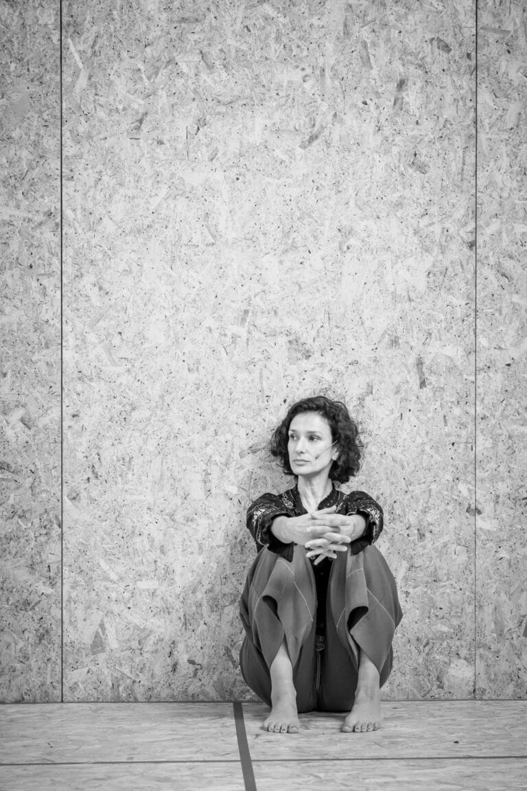 Indira Varma: Sitting on the floor, hugging her knees with her legs bent and her…