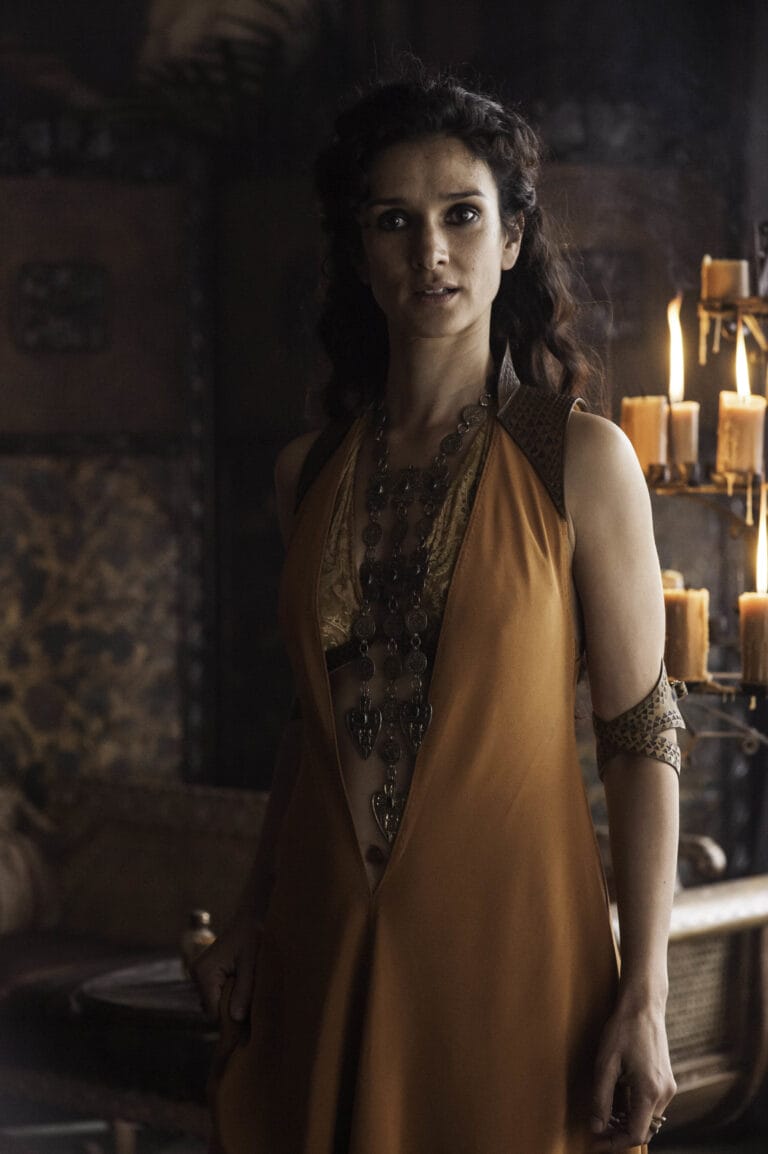 Indira Varma: Wears a long orange dress with a deep neckline and gold details, highlighting…