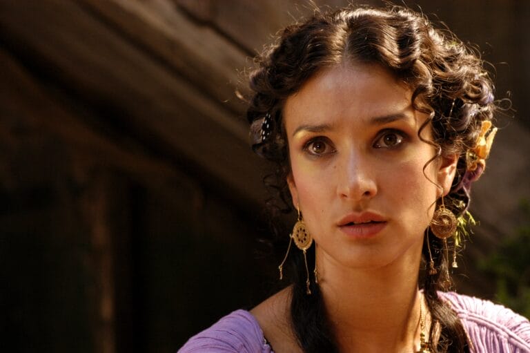 Indira Varma: Wearing a purple dress with shoulder details, in an introspective pose that…
