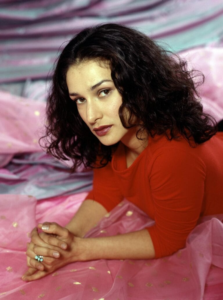 Indira Varma: Wears a long-sleeved red blouse, reclining against a backdrop of pink fabrics…