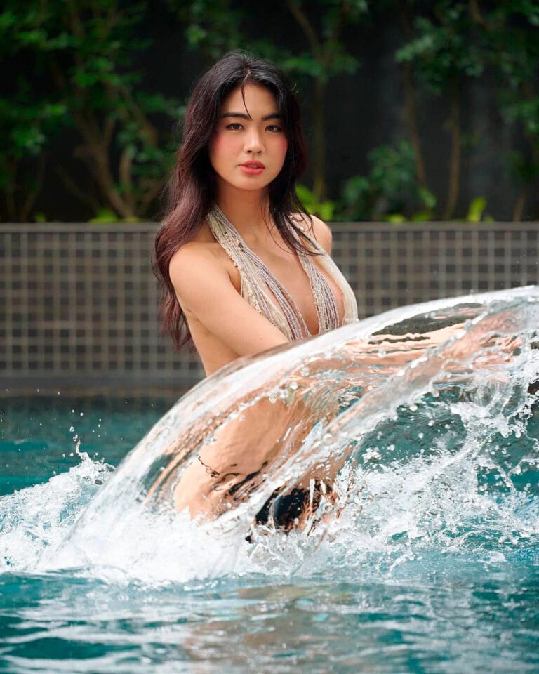 Azi Acosta: Wears a strappy swimsuit that highlights her figure while she plays…