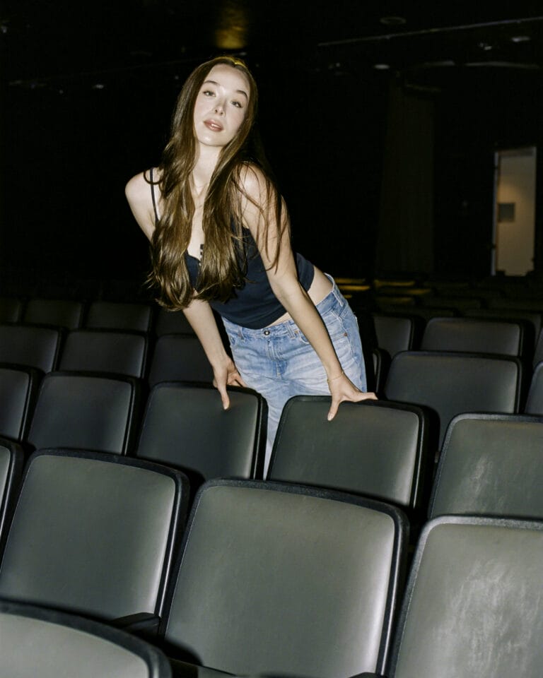 Ella Beatty: Wears a black sleeveless tee and fitted jeans, leaning on the seats of…