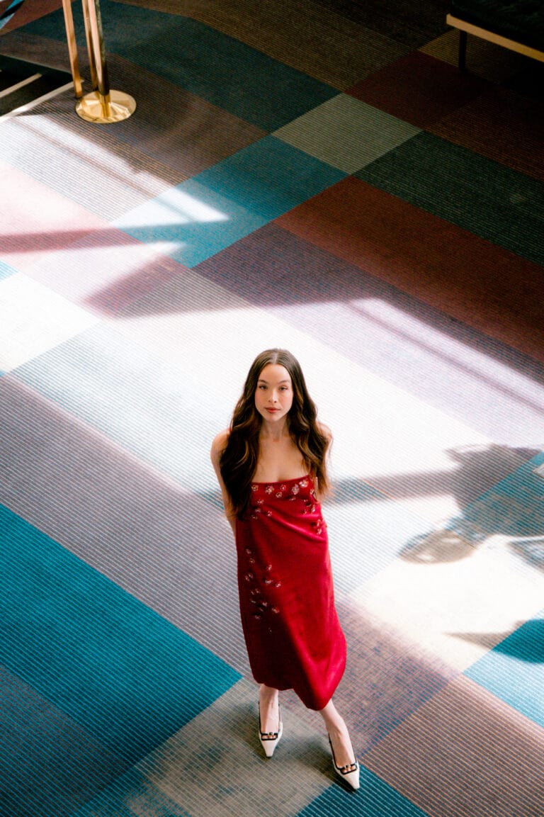 Ella Beatty: Wears a long red dress with delicate embroidery, posing standing with a…